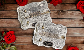 Two custom belt buckles, from Cowboss Silversmiths, on a rustic wooden surface with red roses around